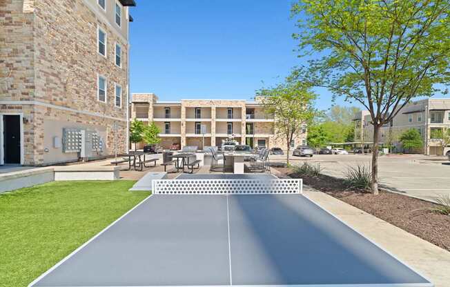 A shiny, metallic ping pong table is in the foreground of a sunny courtyard.