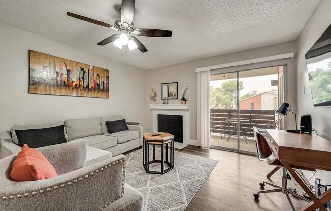 A cozy living room featuring a light gray sectional sofa, a decorative armchair with nailhead trim, a wooden coffee table, and a textured area rug. There is a fireplace with a modern artwork above it, and a sliding glass door leading to a balcony with outdoor views. A small desk is positioned near the window.