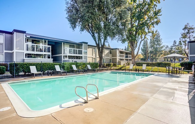 A swimming pool surrounded by trees and chairs in front of apartment buildings.