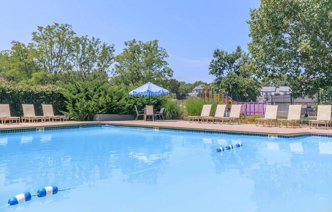 A clear blue swimming pool surrounded by lounge chairs and greenery. In the background, there is a colorful play structure and a shaded seating area with an umbrella. The sky is bright and sunny, indicating a pleasant outdoor atmosphere.