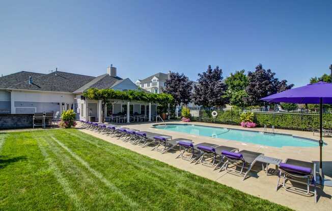 View of pool with purple lounge chairs and lush green grass surrounding. Clubhouse with greenery covered deck can be seen in background.