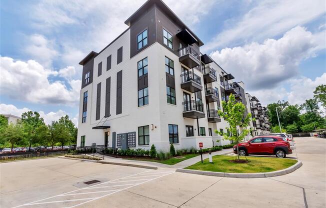 A modern four-story apartment building with balconies near a parking lot with cars and trees