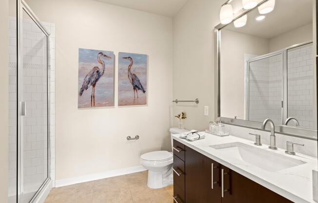 View into the bathrom with a glass enclosed shower and well-lit vanity area with a large mirror at Sylvan Uptown, Denver, CO