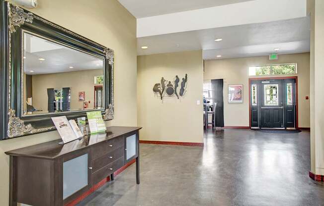 A welcoming community entry here at Allegro featuring a spacious lobby with polished concrete floors, a dark wood console table topped with informational displays, and an ornate framed mirror, complemented by soft recessed lighting, wall art accents, and glass entry doors that allow natural light to flow through the space.