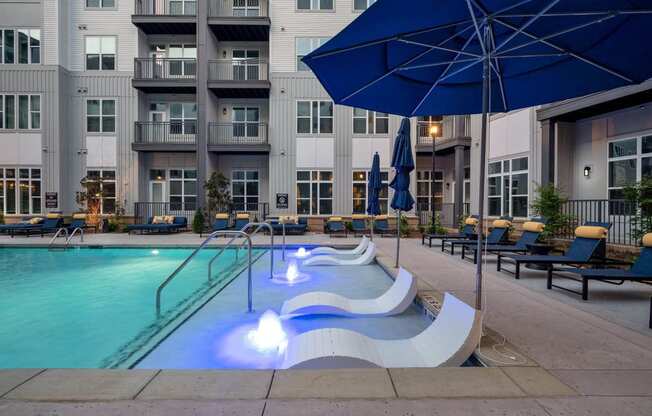 a swimming pool with a blue umbrella and some chairs in front of an apartment building