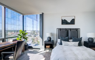 Spacious bedroom with a king-sized bed, crisp white linens, and contrasting black accent pillows, complete with a work desk overlooking an expansive urban landscape.