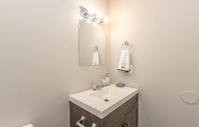 A small bathroom featuring a modern sink with a white countertop, a wall-mounted mirror, and a set of light fixtures above. A towel hangs on a ring by the sink, and there are a few decorative items on the countertop. The walls are painted in a light color, creating a clean, minimalistic aesthetic.