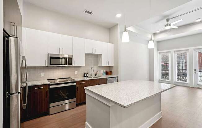 A modern kitchen with stainless steel appliances, a large island in the center, super high ceilings, floor-to-ceiling windows, a patio, and amazing natural light.