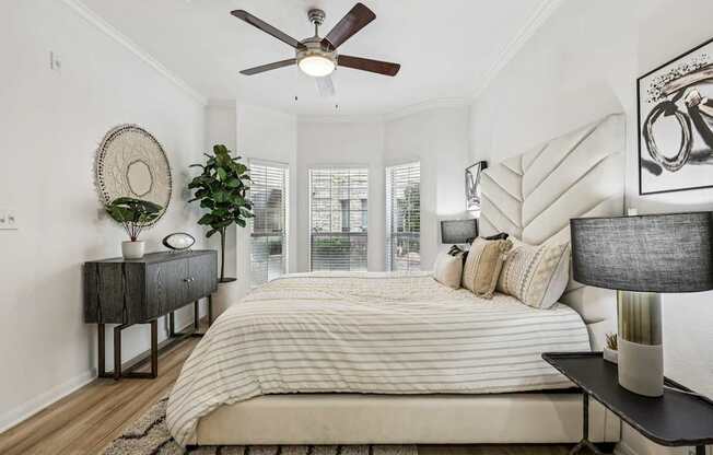 A bedroom with a large bed, a ceiling fan, and a side table with a plant.