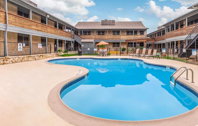 A clear blue swimming pool surrounded by sun loungers and an umbrella, with two-story buildings in the background. The area is well-maintained with green grass and a stone border along the pool. The sky is partly cloudy.