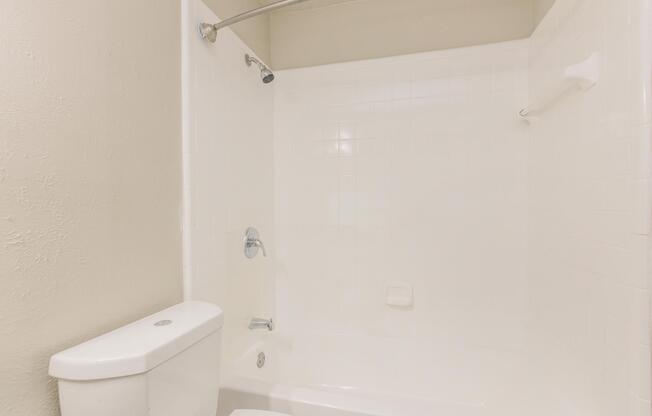 A clean, modern bathroom featuring a white bathtub with a shower, a chrome showerhead, and a white toilet. The walls are tiled in white, creating a bright and airy atmosphere. The space is well-lit and uncluttered, emphasizing a minimalist design.