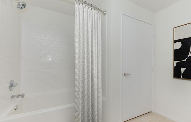 A clean, modern bathroom featuring a white bathtub with a shower, a light-colored shower curtain, and a plain white door. A piece of abstract artwork is displayed on the wall, adding a touch of style to the minimalist decor. The overall atmosphere is bright and inviting.
