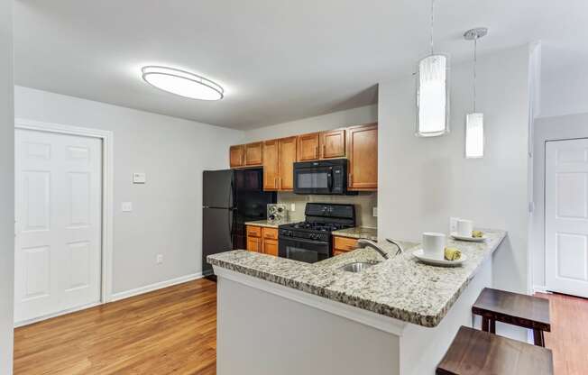 Apartments For Rent In Aurora, IL - The Apartments At Kirkland Crossing - Kitchen With Granite-Style Countertops, Wood-Style Flooring, Brown Cabinets, And Black Appliances.