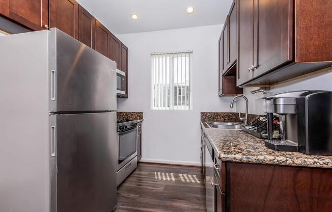 A modern kitchen featuring dark wooden cabinets, stainless steel appliances including a refrigerator, oven, and microwave. The countertops are made of granite. A small window with vertical blinds allows natural light to enter the space, and a coffee maker is placed on the counter. The flooring is dark and the overall design is contemporary.