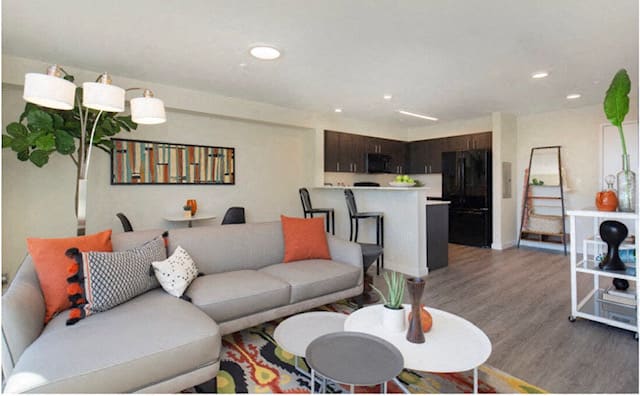 Living Room with Plank Flooring, Cozy Grey Couch, Nesting Tables, and Curved Lamp with Kitchen View at Arabella Apartment Homes, Washington