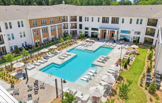 A large swimming pool surrounded by lounge chairs and umbrellas in front of a multi-story apartment building.