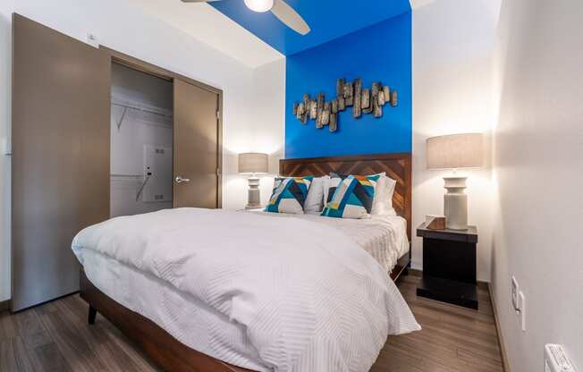 Bedroom with Closet, White Comforter Bedding, Tan Double Door, Hardwood Inspired Floor, Small Bedside Table with Lamp On Top