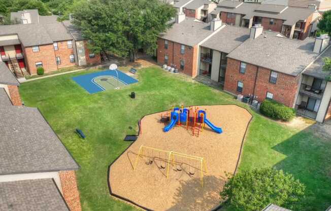 A playground with a slide, swings, and a basketball hoop in a grassy area surrounded by brick buildings.