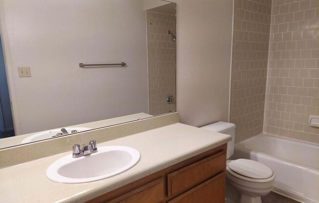 A clean bathroom featuring a white sink with a chrome faucet, a large mirror above the sink, a beige tiled wall, a bathtub with a showerhead, and a white toilet. The floor is covered with neutral-colored tiles, and there is a towel rack on the wall.