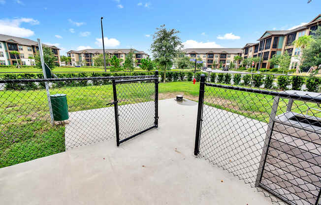 a fenced in dog park with trash cans and apartment buildings in the background
