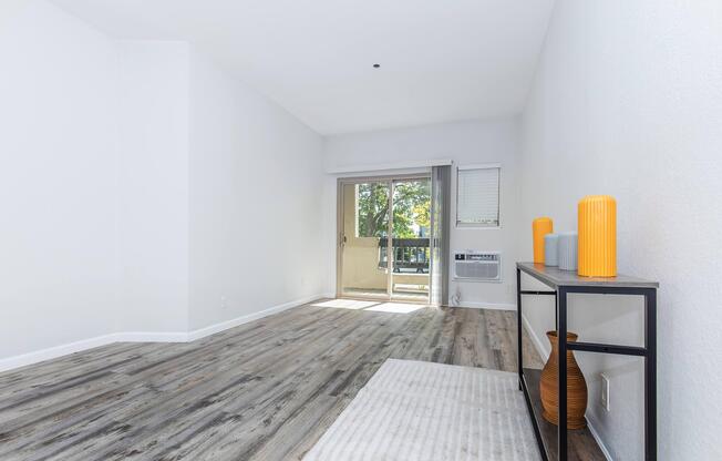 Bright, spacious interior of a living room featuring light wood-like flooring, white walls, and a sliding glass door leading to a balcony. A modern console table with orange decorative vases is positioned against the wall. Natural light fills the room, creating an inviting atmosphere.