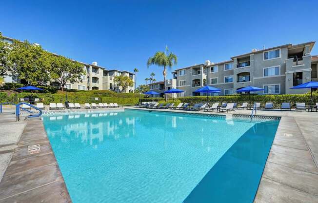 A large swimming pool in front of apartment buildings with sun loungers and umbrellas.