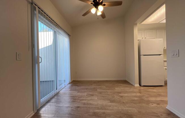 An empty room with wooden flooring, a ceiling fan, and a large sliding glass door leading to a balcony. A white refrigerator is visible in the corner, and the walls are painted a light color, creating a bright and airy atmosphere.