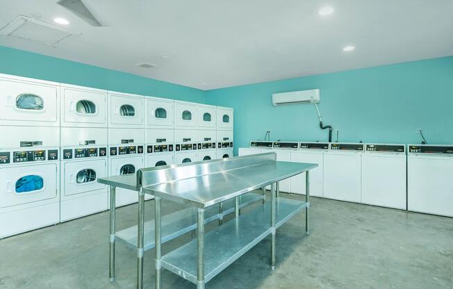 A clean and bright laundry room featuring several white washing machines and dryers against a light blue wall. In the center, there is a stainless steel folding table. LED lights illuminate the space, enhancing its modern and tidy appearance. An air conditioning unit is mounted on the wall.