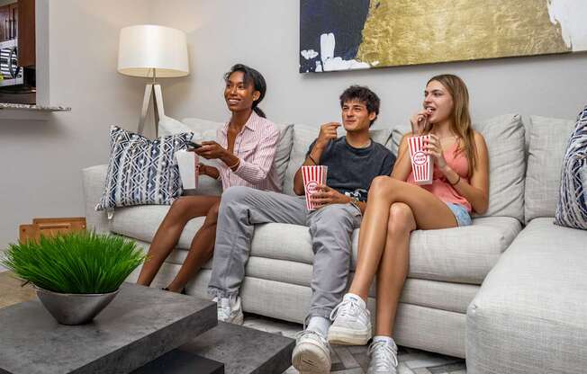 Three people are sitting on a couch with popcorn and a remote control.