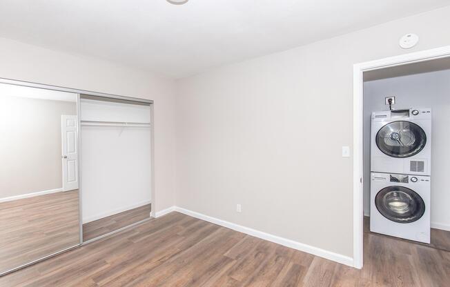 A well-lit room with light-colored walls and wooden flooring. It features a mirrored sliding closet door on the left, and on the right, there is a stacked washer and dryer. The space is empty, promoting a clean and spacious feel.