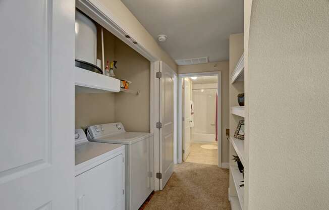 A convenient in-home laundry area here at Tuscany at Faudree with a full-size washer and dryer tucked into a hallway closet. Open shelving above provides storage for supplies, while plush carpet, neutral walls, and nearby built-in display shelves create a clean, functional transition space leading toward the bathroom.
