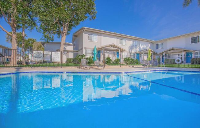 A clear blue swimming pool surrounded by lounge chairs and umbrellas, with well-maintained greenery in the background. White buildings with multiple windows are visible beyond the pool area under a bright blue sky. The scene conveys a relaxing, inviting atmosphere.
