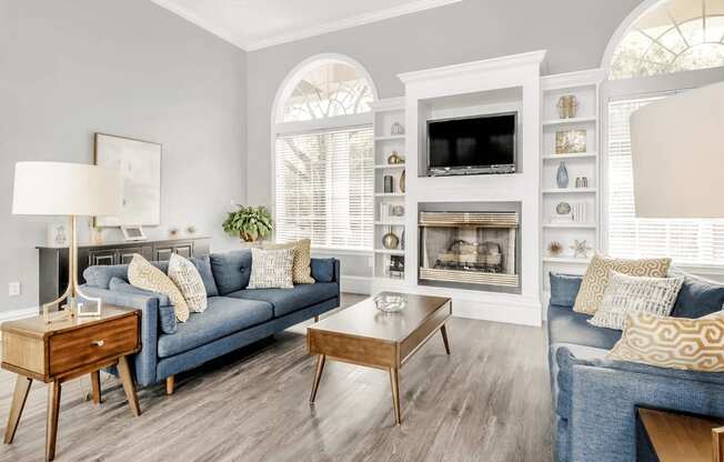 Clubhouse interior with blue couches, brown accent tables, fireplace, wall mounted television, gray walls with large windows