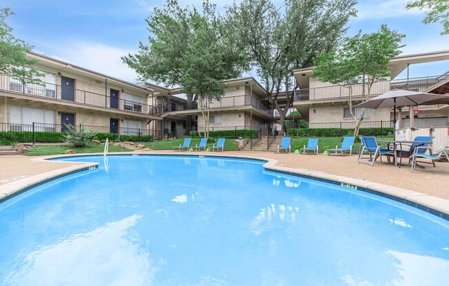A clear blue swimming pool surrounded by lounge chairs and umbrellas, with a landscaped area featuring trees and shrubs. In the background, there are two-story apartment buildings with balconies. The scene is bright and inviting, perfect for relaxation and leisure.