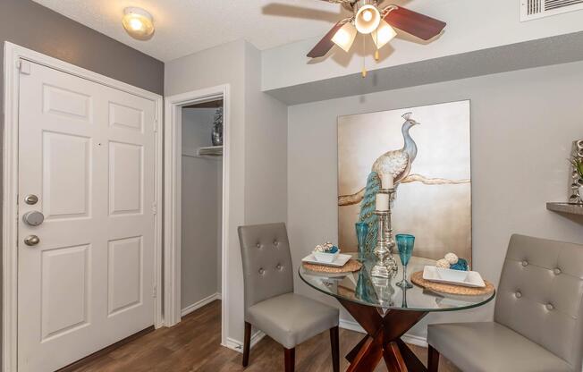 A cozy dining area featuring a round glass table with two gray upholstered chairs. The table is set with decorative plates and blue glasses. A peacock-themed artwork adorns the wall, adding a touch of elegance. Soft lighting from a ceiling fan enhances the warm atmosphere. A door and closet are visible in the background.