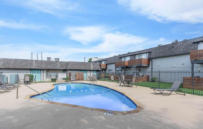 Swimming pool surrounded by lounge chairs and a gated area. There are two multi-story buildings in the background with balconies and a clear blue sky above. The pool has a curved shape and is well-maintained, providing a relaxing atmosphere.