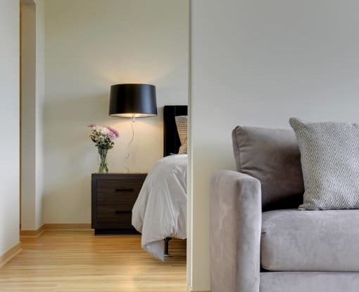 Flat J living area with grey couch and glimpse into bedroom behind couch with black nightstand and lit lamp