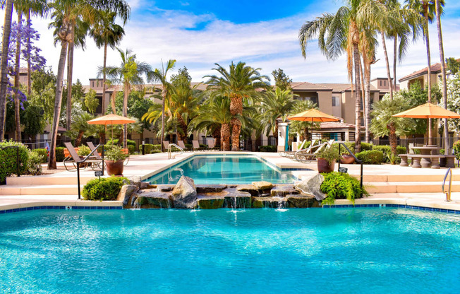 Crystal clear blue lap pool flowing over water feature into a resort-style pool surround by chaise lounge chairs, palm trees and plants