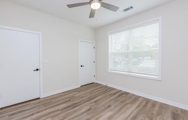 A brightly lit room featuring light-colored walls and a light wood floor. There is a ceiling fan with blades, a window with blinds allowing natural light, and two doorsâone is a standard door and the other appears to be a closet door. The space is minimalistic and uncluttered.