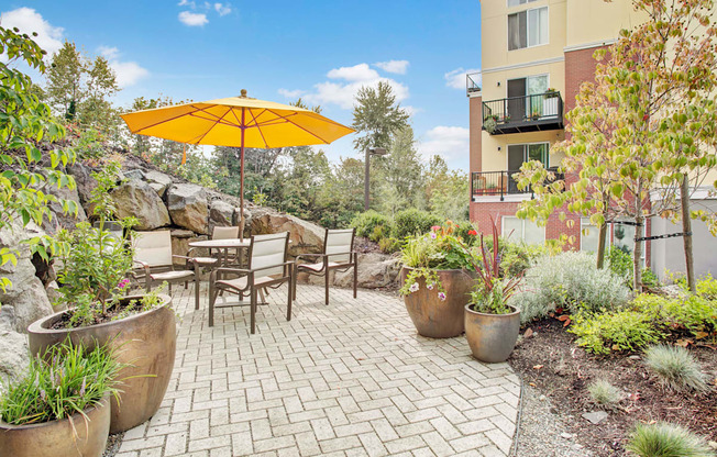 A landscaped outdoor courtyard here at Heritage Hills featuring a paved patio with café-style seating arranged beneath a bright yellow umbrella. The space is surrounded by large decorative planters, lush greenery, and natural stone accents, with nearby apartment balconies and mature trees creating a peaceful setting for outdoor relaxation and casual gatherings.