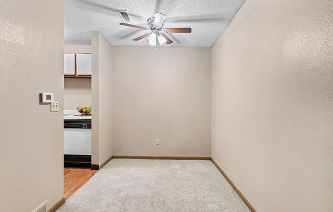 an empty room with a ceiling fan and a kitchen in the background