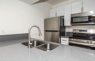 A modern kitchen featuring a gray countertop with a stainless steel double sink, a sleek faucet, white cabinetry, and stainless steel appliances including an oven, microwave, and refrigerator. The walls are painted a light color, creating a bright and inviting atmosphere.