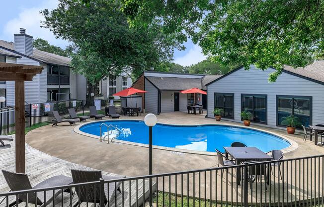 A serene outdoor pool area surrounded by lounge chairs and a wooden deck, with red umbrellas providing shade. Nearby, there are tables and chairs for relaxation, and two buildings with gray siding are visible in the background, set against a green landscape of trees.