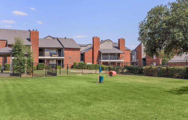 A well-maintained grassy area with a fence, surrounded by brick apartment buildings. In the foreground, there is a playground structure and a large red tube. The sky is clear with a few clouds, and trees are visible in the background, enhancing the outdoor space.