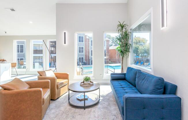 A modern lounge space featuring two brown leather armchairs and a blue fabric sofa. A round coffee table sits in the center, surrounded by light-colored walls and large windows that overlook an outdoor pool area. Natural light fills the room, creating a bright and inviting atmosphere.