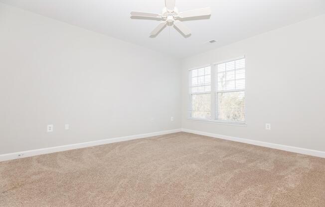 Empty room with light gray walls and beige carpet. A ceiling fan is visible, and there are two windows with white blinds allowing natural light to enter, creating a bright and spacious atmosphere. The room is unfurnished and clean.