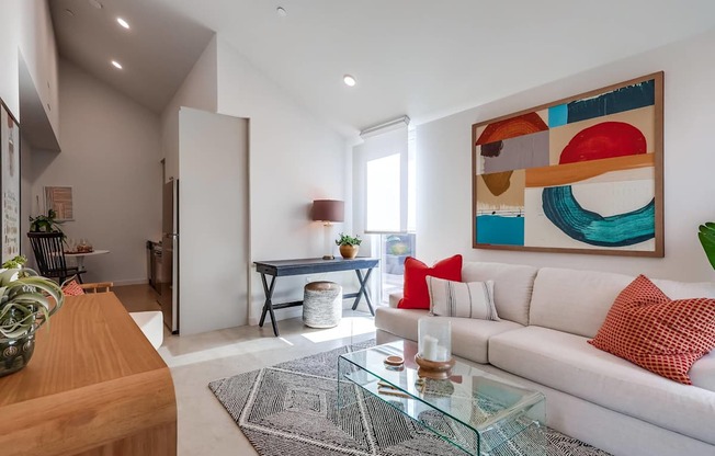 A modern living room with a white sofa, a glass coffee table, and a colorful abstract painting on the wall.