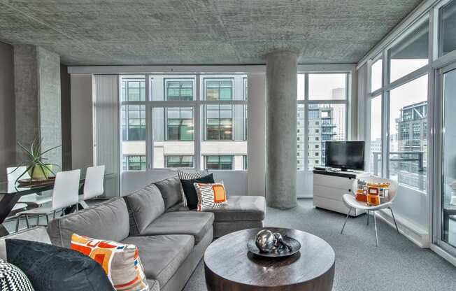 a living room with a gray couch and a round table   and large windows