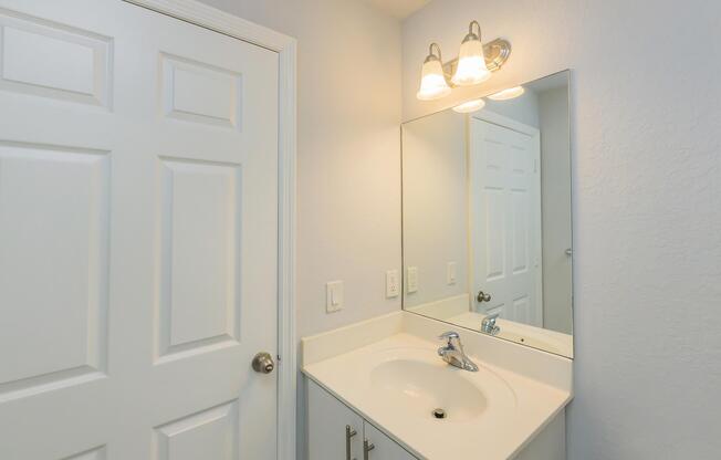 A clean and bright bathroom featuring a white sink with modern faucets, a large wall mirror, and a light fixture above. The light blue walls and white door create a fresh atmosphere. The design is simple yet functional, ideal for everyday use.