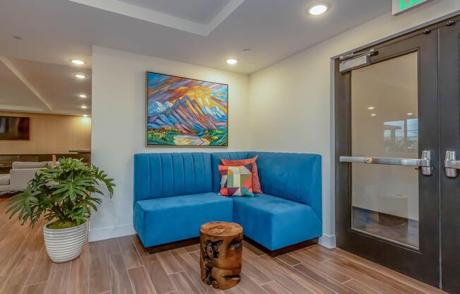 A modern lounge area featuring a blue upholstered couch with a colorful geometric pillow, a wooden stump as a side table, and a potted plant. A vibrant mountain landscape painting hangs on the wall, and a glass door leads to another space, creating a welcoming and stylish atmosphere.